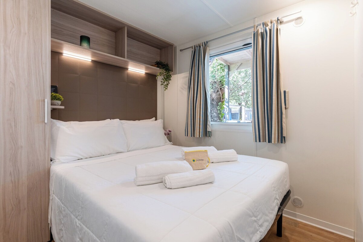 Una cama bien hecha con ropa blanca, una mesita de noche con una planta y una ventana con cortinas a rayas.