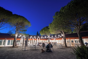 An outdoor dining area with tables and chairs, surrounded by trees, and a building with a red roof in the background. - Club Del Sole Marina Julia Family Collection (Monfalcone)