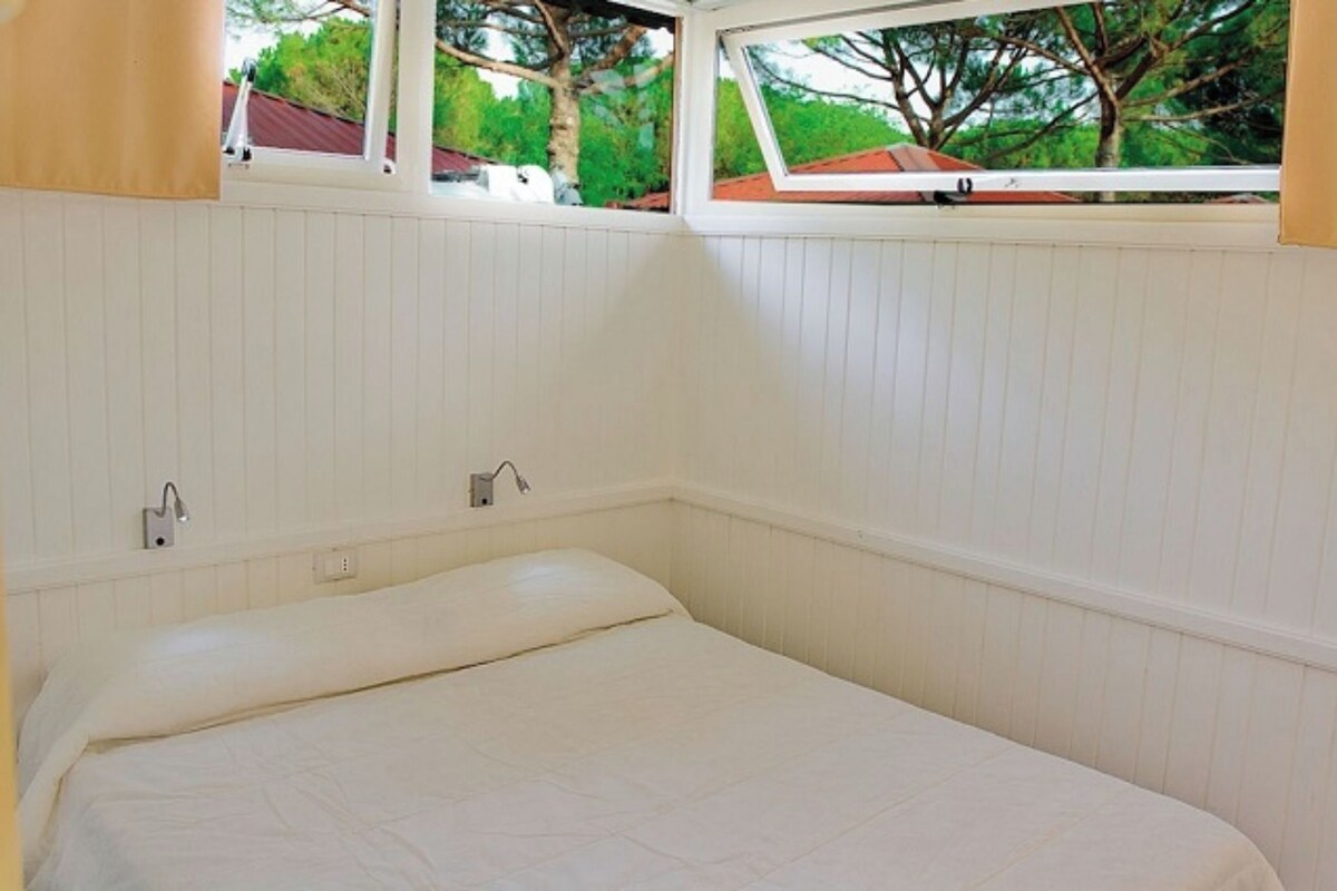 A bedroom with a bed, two wall-mounted light fixtures, and a window with a view of trees and a red-roofed building.