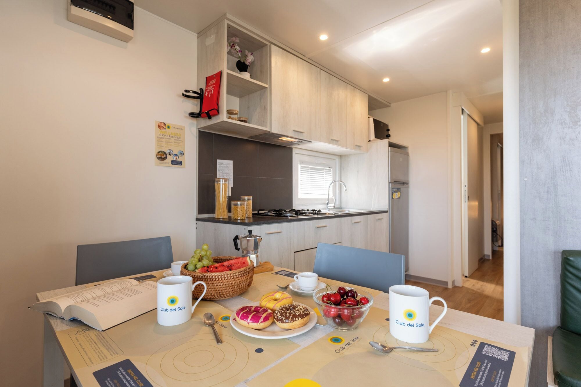 A modern kitchen with white cabinets, a gas stove, and a sink. There is a dining table with a basket of fruit, a plate of pastries, and two mugs labeled 'Club del Sole'.