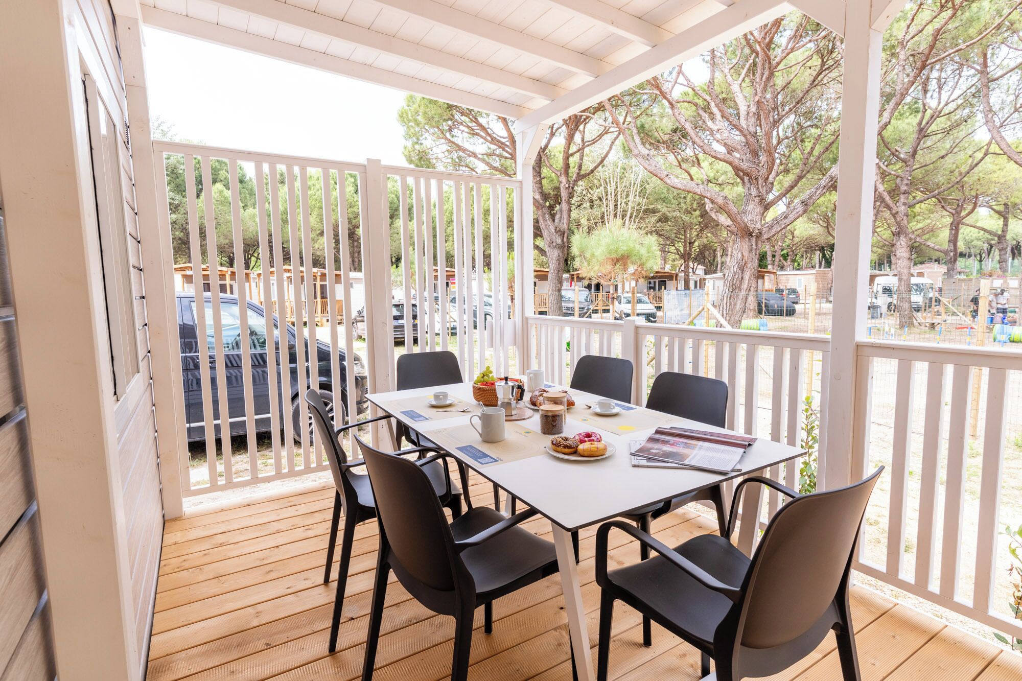 A covered outdoor seating area with a table set for four, surrounded by chairs, overlooking a parking area with trees and parked cars.