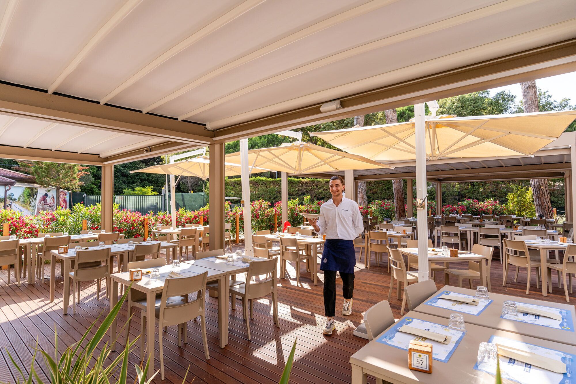 An outdoor dining area with tables and chairs, a waiter, and a covered roof structure.