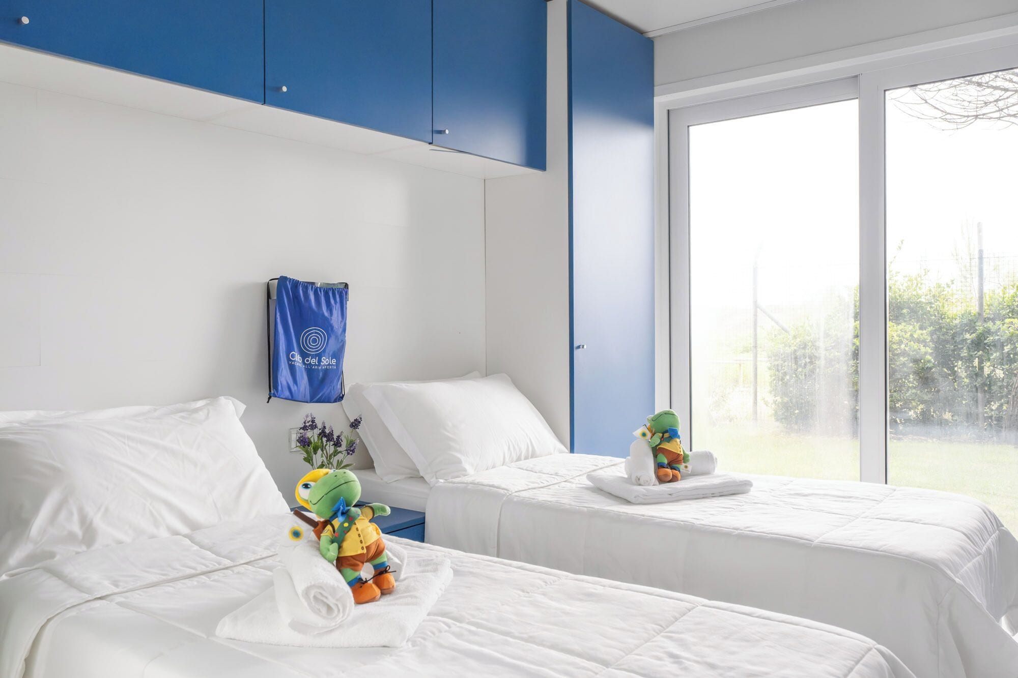 Habitación de hotel con dos camas, una ventana con vistas, un toallero azul y una pequeña mesa con un juguete.
