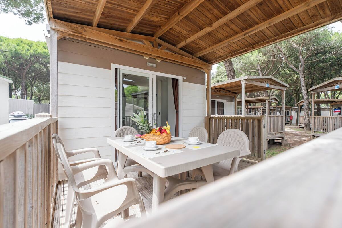 Un espace extérieur abrité, aménagé pour prendre les repas, avec une table dressée pour deux personnes, des chaises et un panier de fruits.