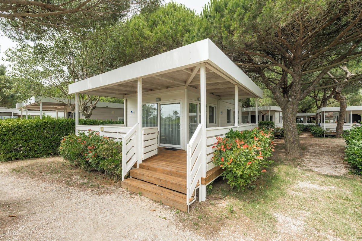 Un espace extérieur abrité, doté d’un escalier en bois menant à une porte, et entouré de verdure et d’arbres.