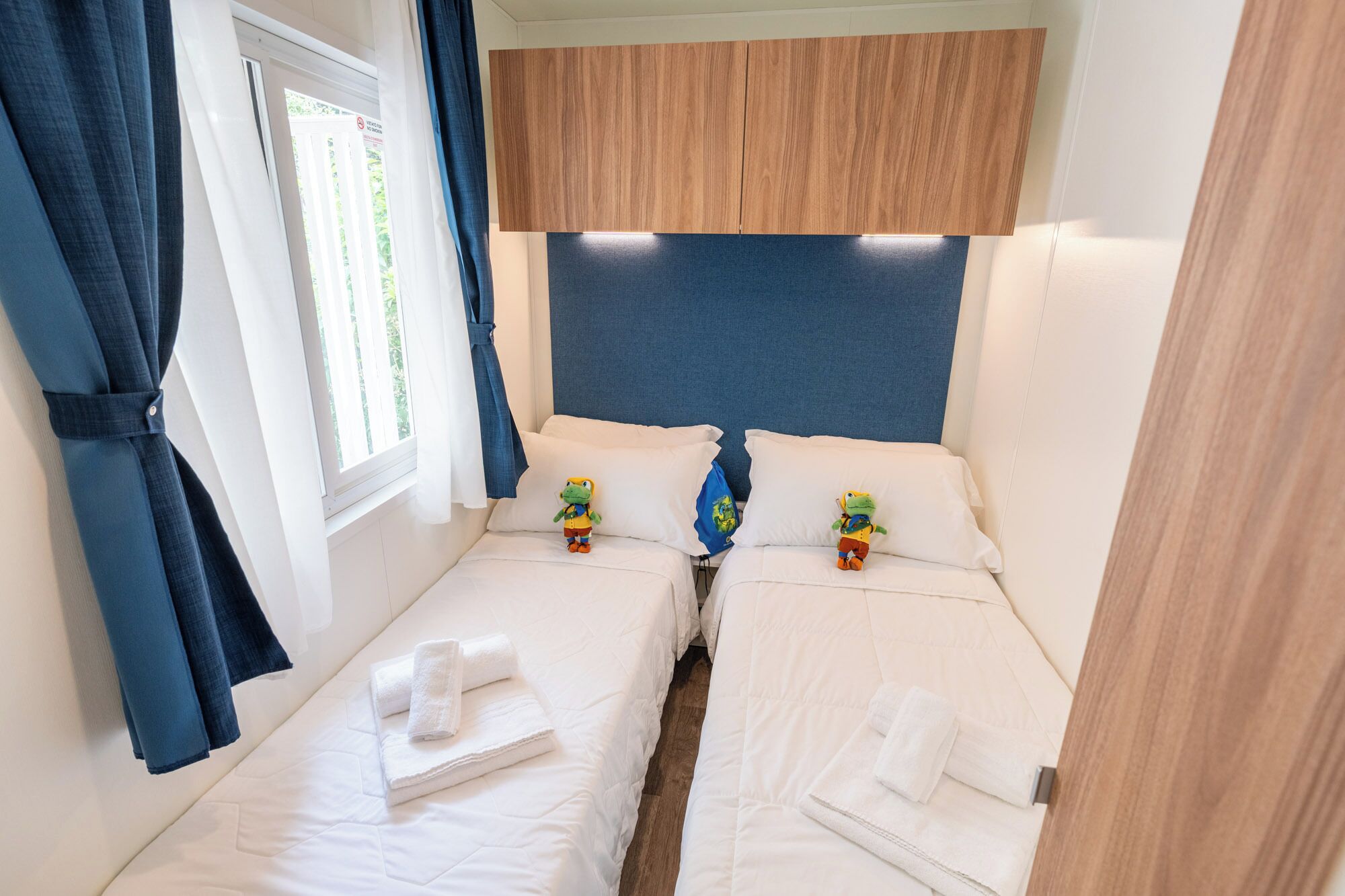 A small, cozy room with two beds, a window with curtains, and a wooden headboard.