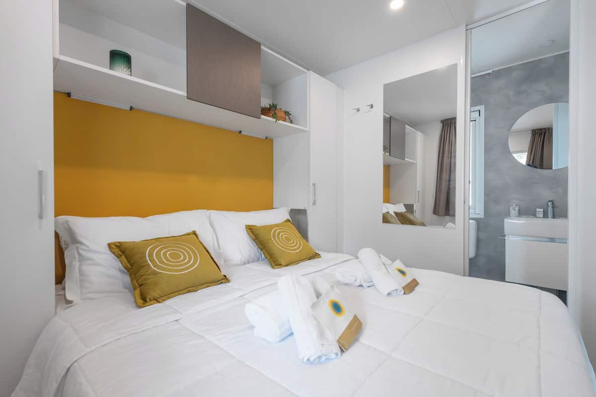 Une chambre d’hôtel avec un lit, du linge de lit blanc, une tête de lit, un miroir et une salle de bain visible à travers une porte ouverte.