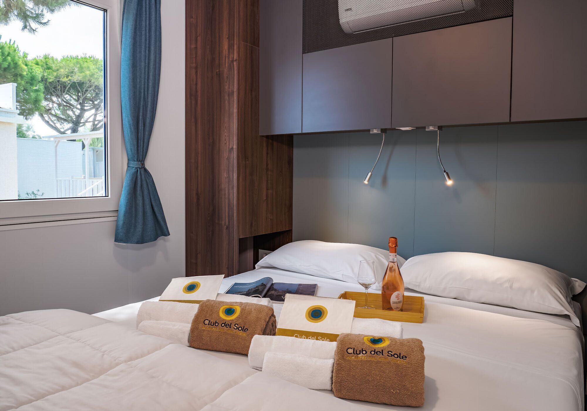 Une chambre d’hôtel avec un lit, des oreillers, une table de chevet avec une bouteille et des verres, une fenêtre avec des rideaux et une vue sur les arbres à l’extérieur.