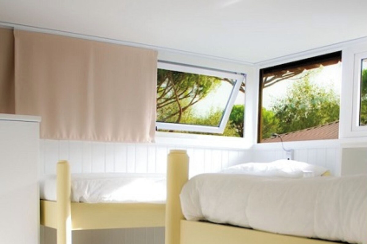 A bunk bed room with a white refrigerator, beige curtains, and windows with a view of trees.