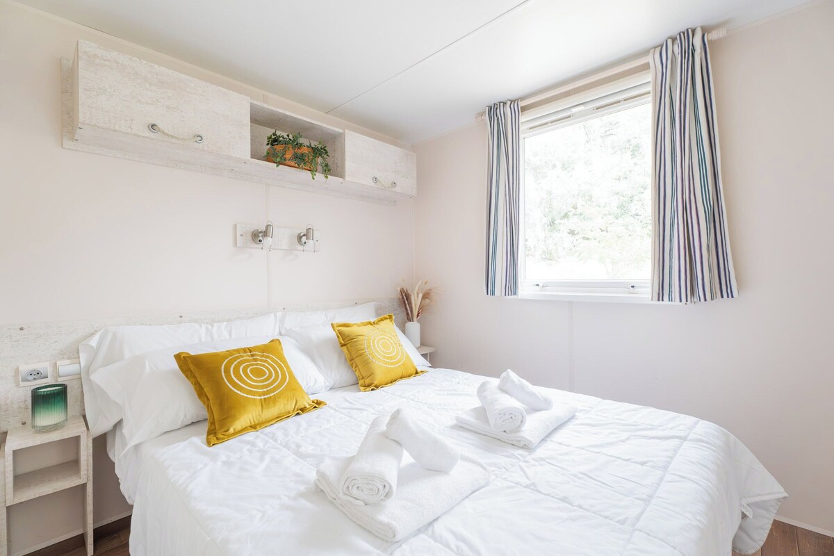 A neatly made bed with white bedding and two yellow pillows, a bedside table with a lamp, and a window with striped curtains.