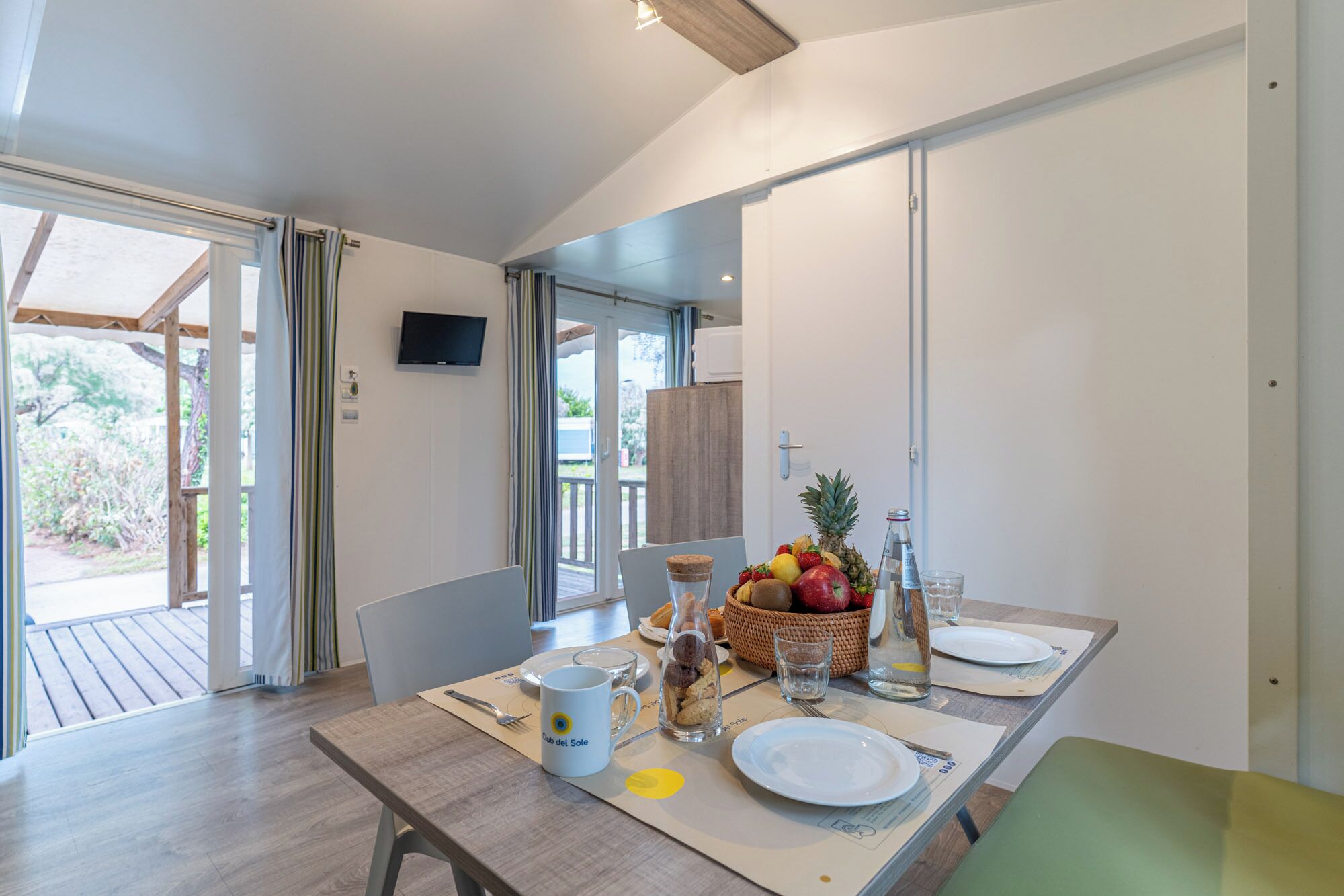 Un área de comedor con una mesa dispuesta para dos, una cesta de frutas y un televisor en la pared.
