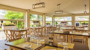 A restaurant with wooden tables and chairs, numbered seating, and large windows with greenery outside. - Club del Sole Vigna sul Mar Family Collection (Comacchio)