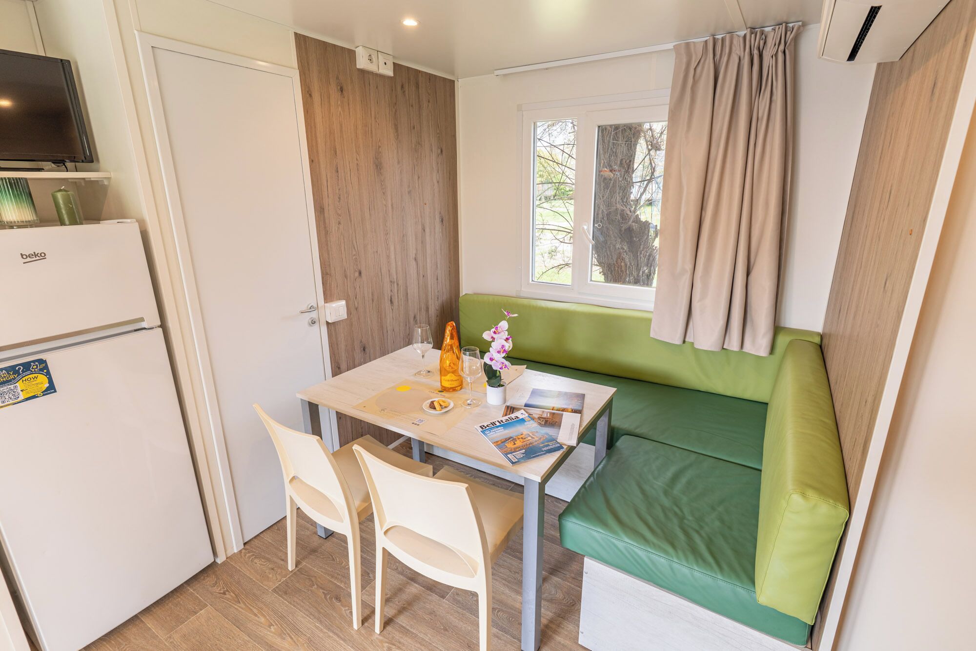 A compact kitchenette with a fridge, a table with chairs, a green cushioned bench, and a window with curtains.