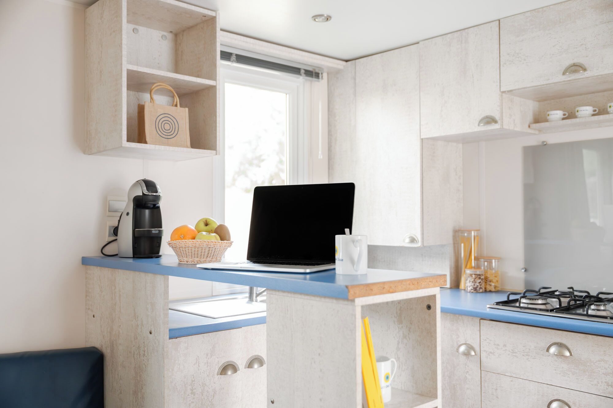 A modern kitchen with a laptop, coffee maker, basket of fruit, and a gas stove.
