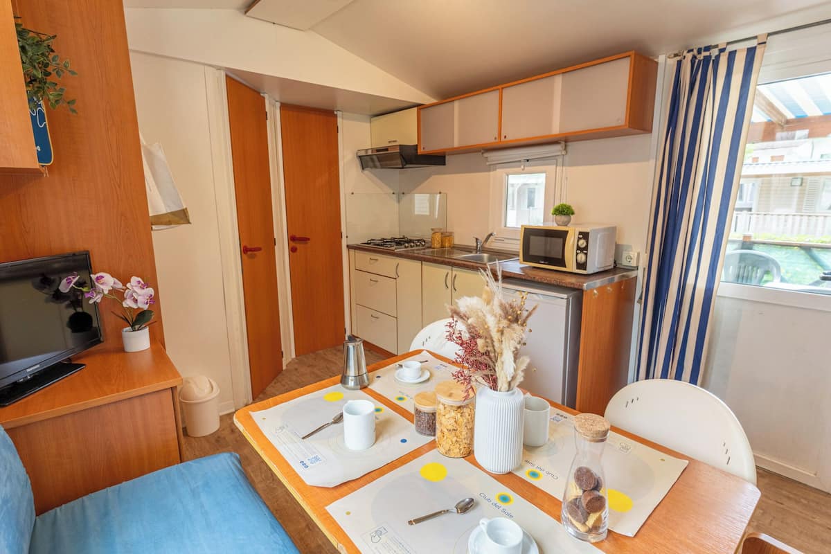 A compact kitchen with a microwave, stove, and sink. There is a dining table set with plates, cutlery, and a vase with dried flowers.