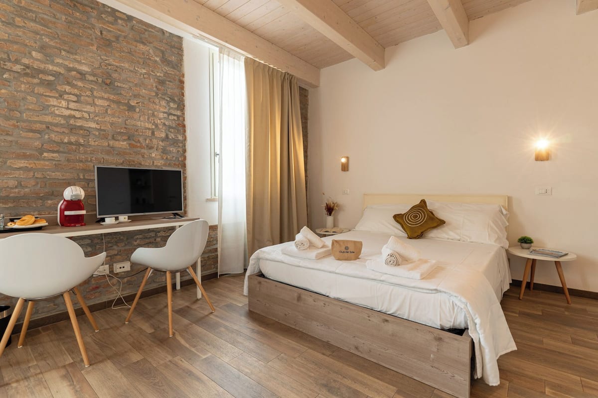 A hotel room with a brick wall, a wooden desk with a TV, a bed with white linens, and a small bedside table.