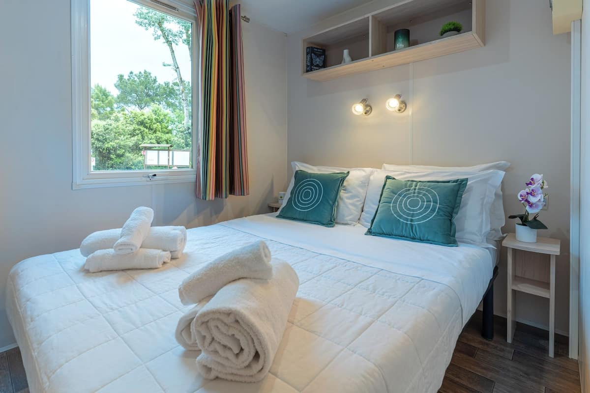 A neatly made bed with white bedding, a window with curtains, and a bedside table with a plant.