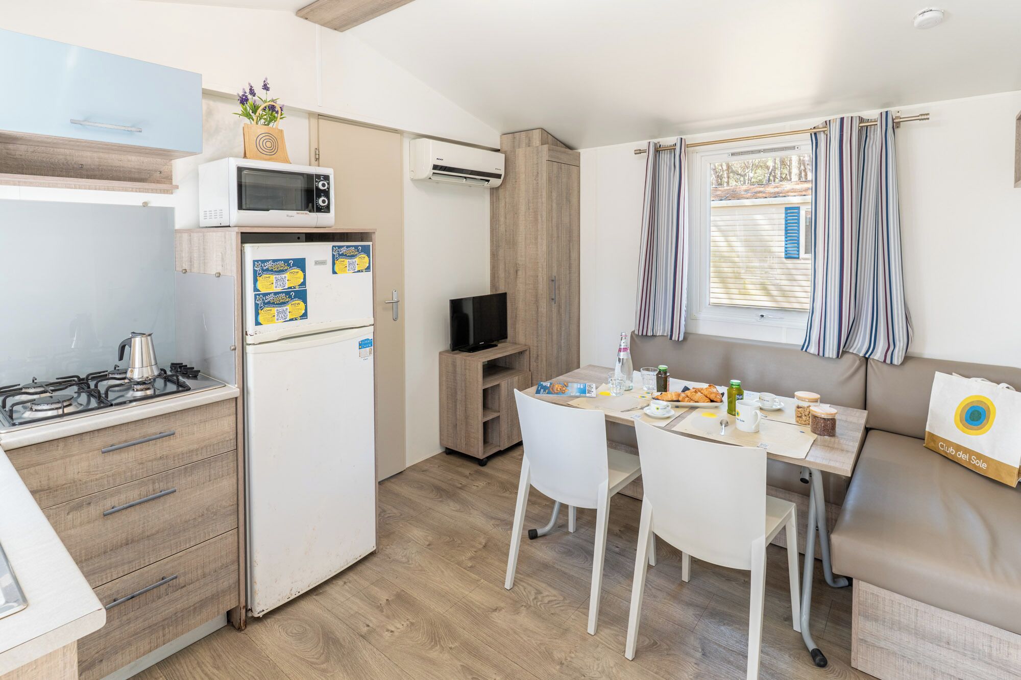 A compact kitchen with a gas stove, microwave, and refrigerator. There's a dining area with a table and chairs, a television, and a window with curtains.