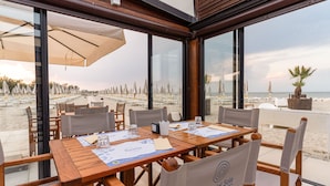 An outdoor dining area with tables and chairs, large windows, and a view of the beach. - Club Del Sole Spina Family Collection (Comacchio)