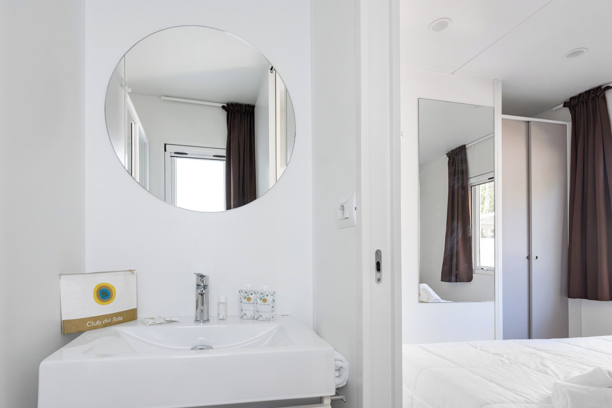 Un bagno d'hotel con un lavandino bianco, uno specchio rotondo e un asciugamano appeso al muro.