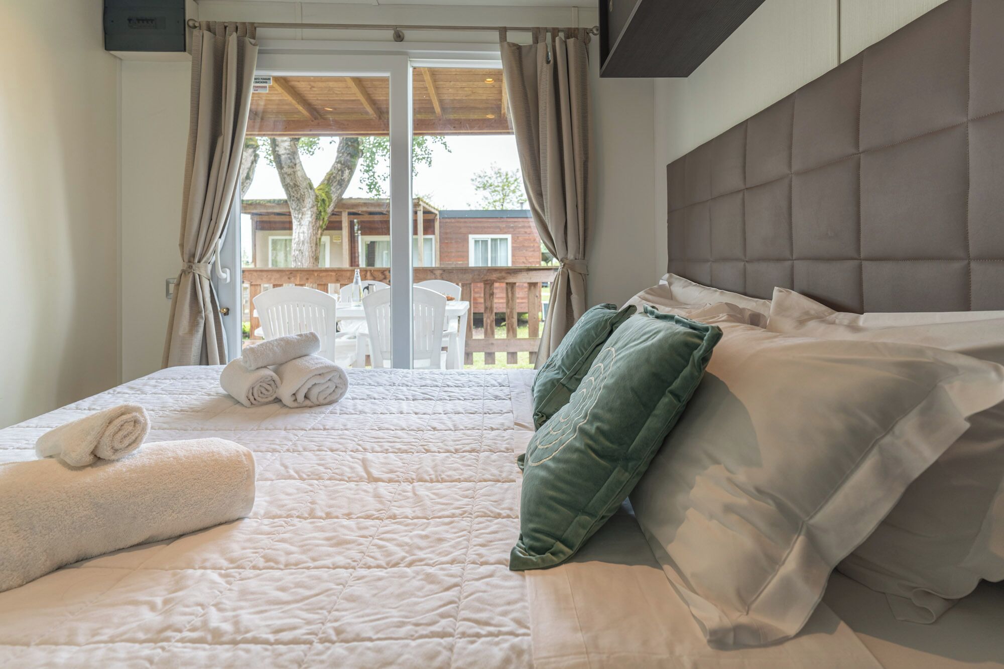 A bedroom with a bed, pillows, a towel, and a view of an outdoor area with a wooden deck and chairs.