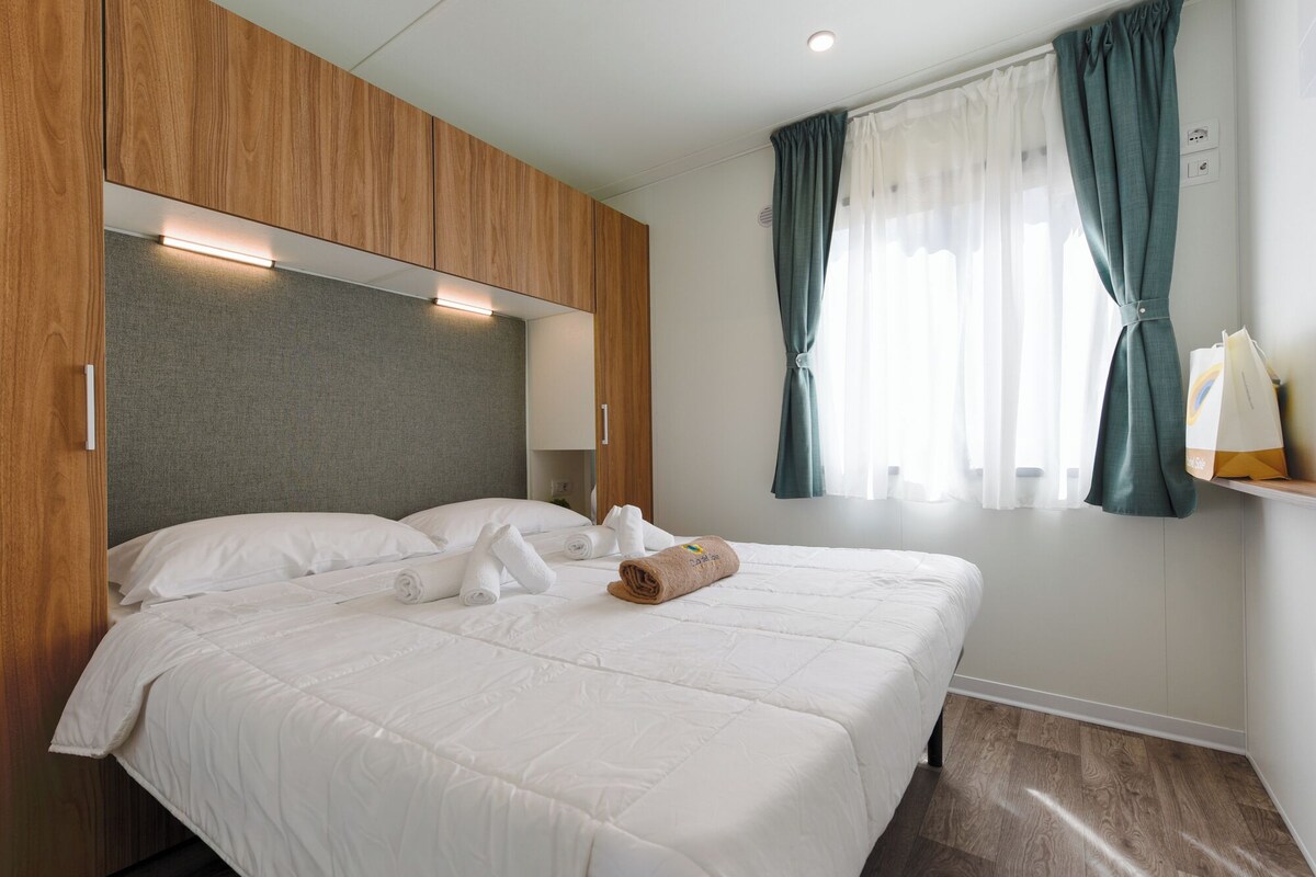 A hotel room with a wooden headboard, a bed with white bedding, a window with curtains, and a towel on the bed.