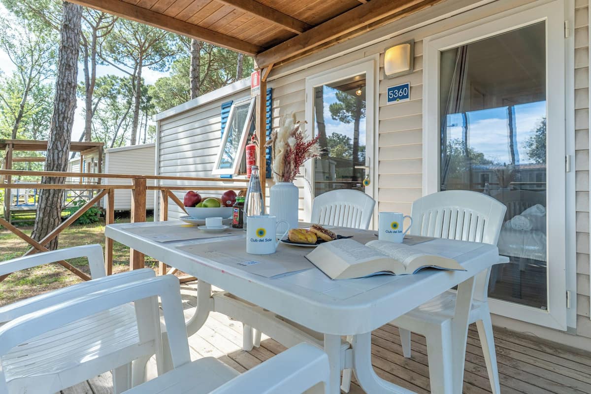 A covered outdoor seating area with a table set for two, a chair, and a window with a view of trees.