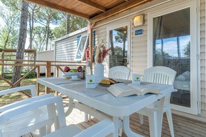 A covered outdoor seating area with a table set for two, a chair, and a window with a view of trees. - Club Del Sole Spina Family Collection (Comacchio)