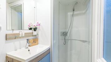 Un baño con mampara de vidrio, un lavamanos blanco con tocador de madera, un espejo con repisa superior y una puerta azul.