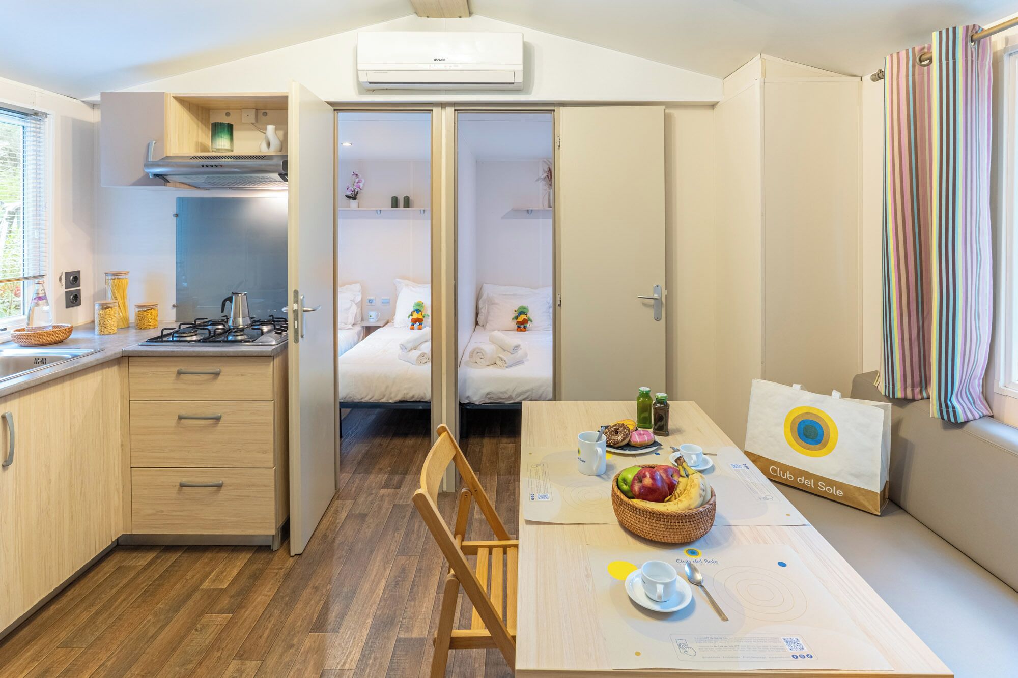 Ein gemütlicher Raum mit Küchenzeile, einem Bett mit hochwertiger Bettwäsche, einem Esstisch mit Frühstücksangeboten und einem Fenster mit gestreifteren Vorhängen.