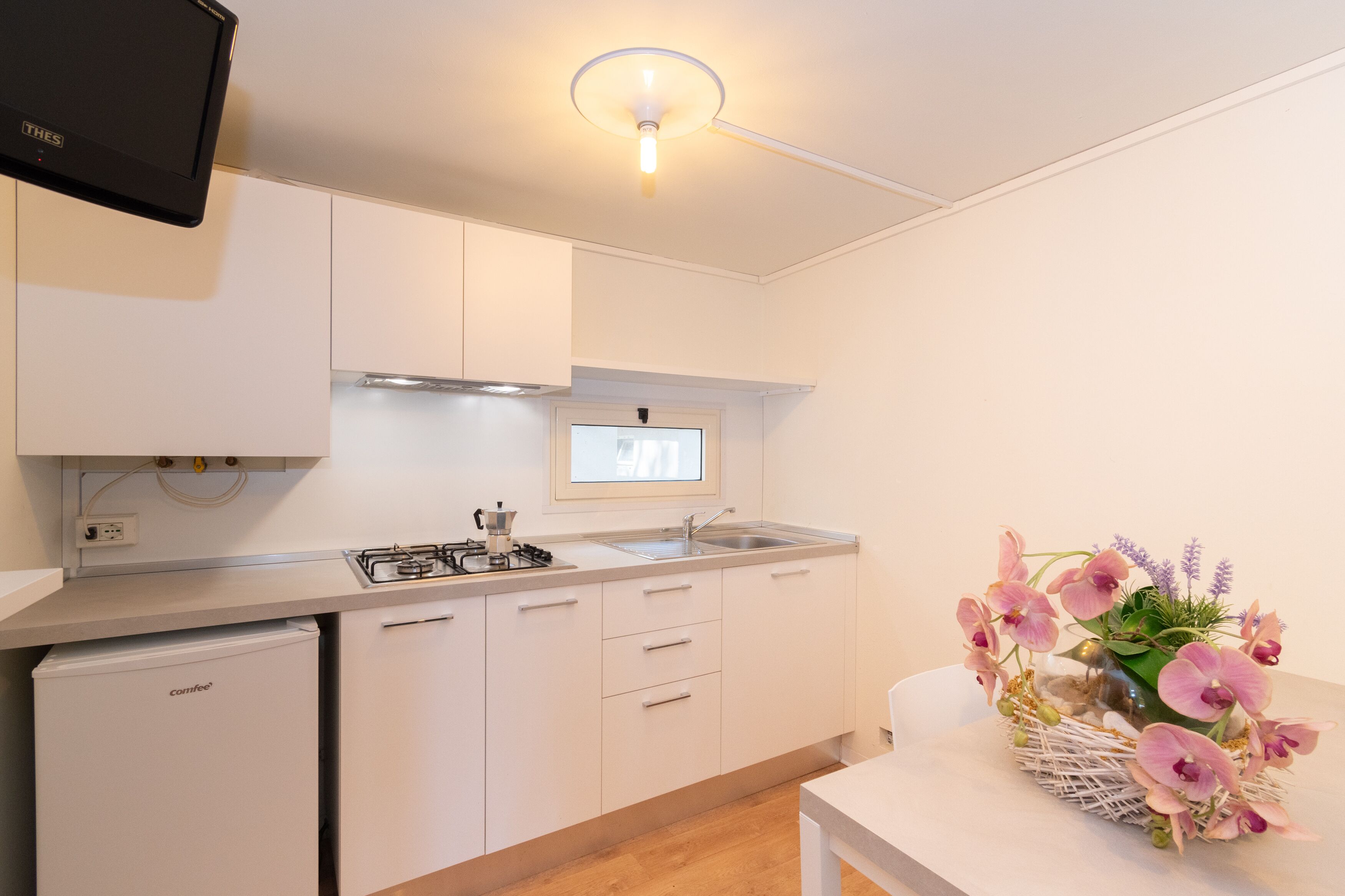 Une cuisine moderne avec des armoires blanches, une cuisinière encastrée et un évier. On y trouve un petit réfrigérateur et un arrangement décoratif d’orchidées sur le comptoir.