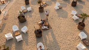 A beachside lounge area with white lounge chairs, wooden tables, and potted plants. - Club Del Sole Spina Family Collection (Comacchio)