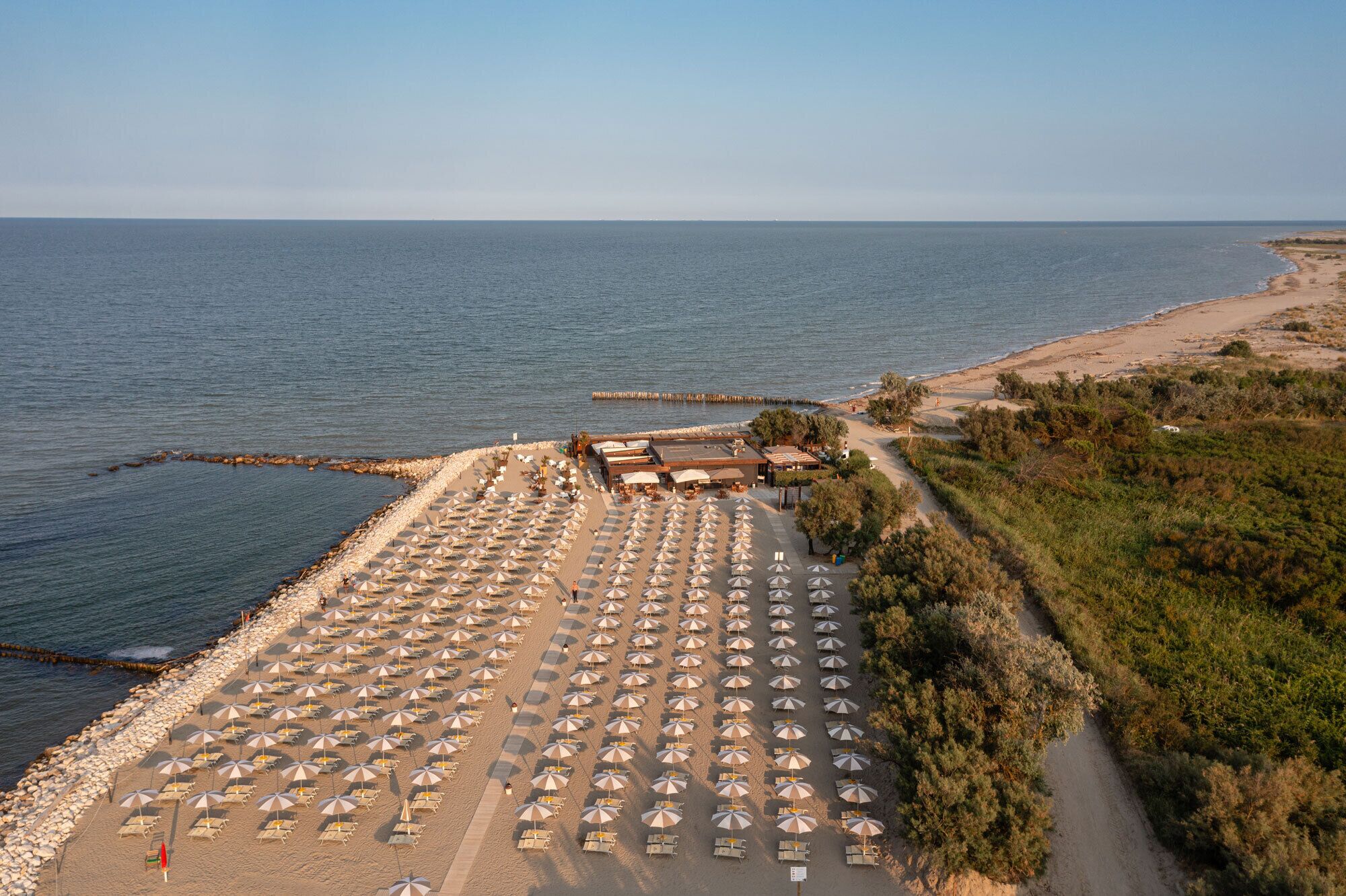 An aerial view of a beachside hotel with rows of sunbeds and umbrellas, a rocky shoreline, and a clear view of the sea.
