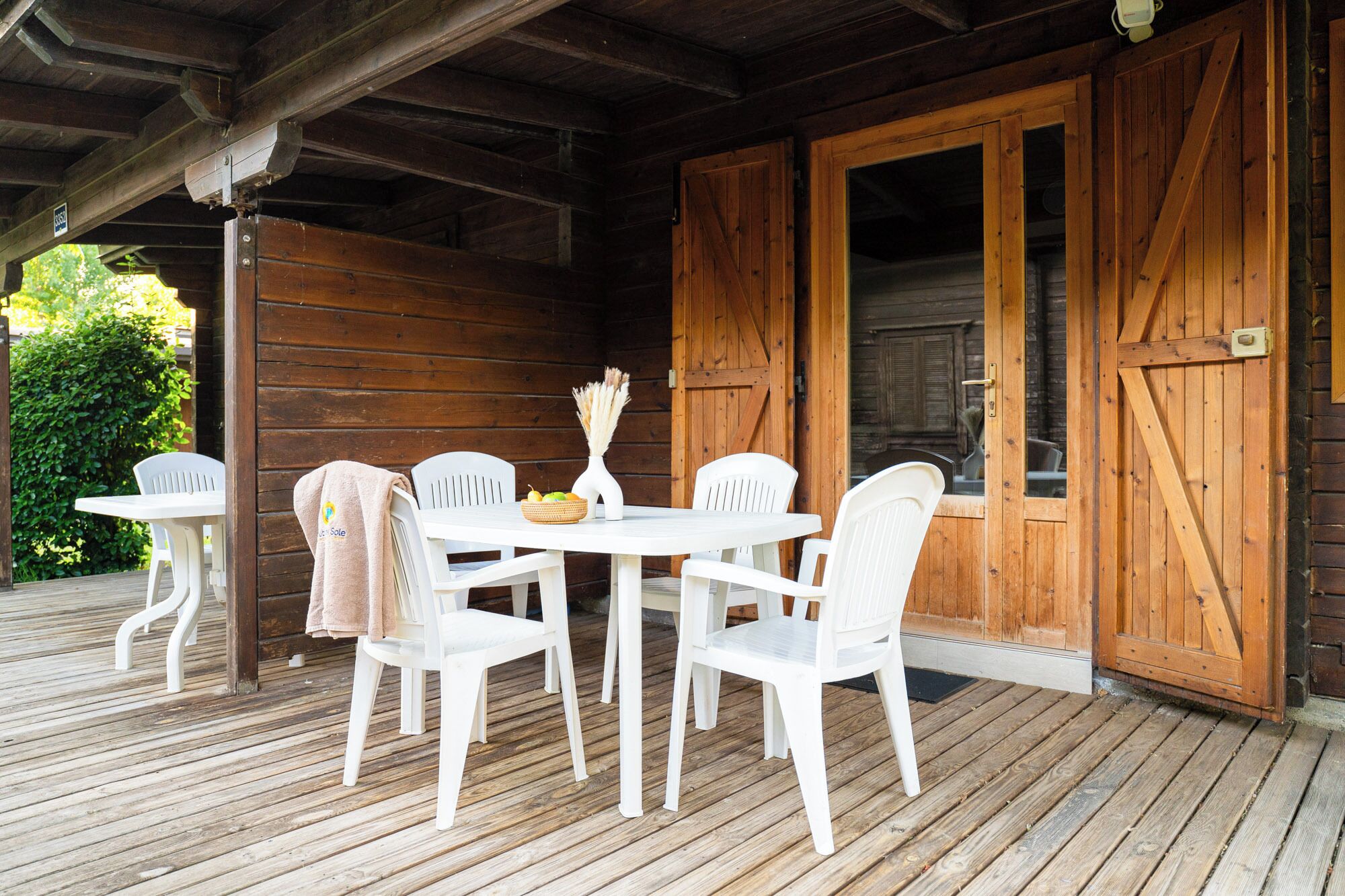 A wooden deck with a table and chairs, a wooden door, and a view of greenery.