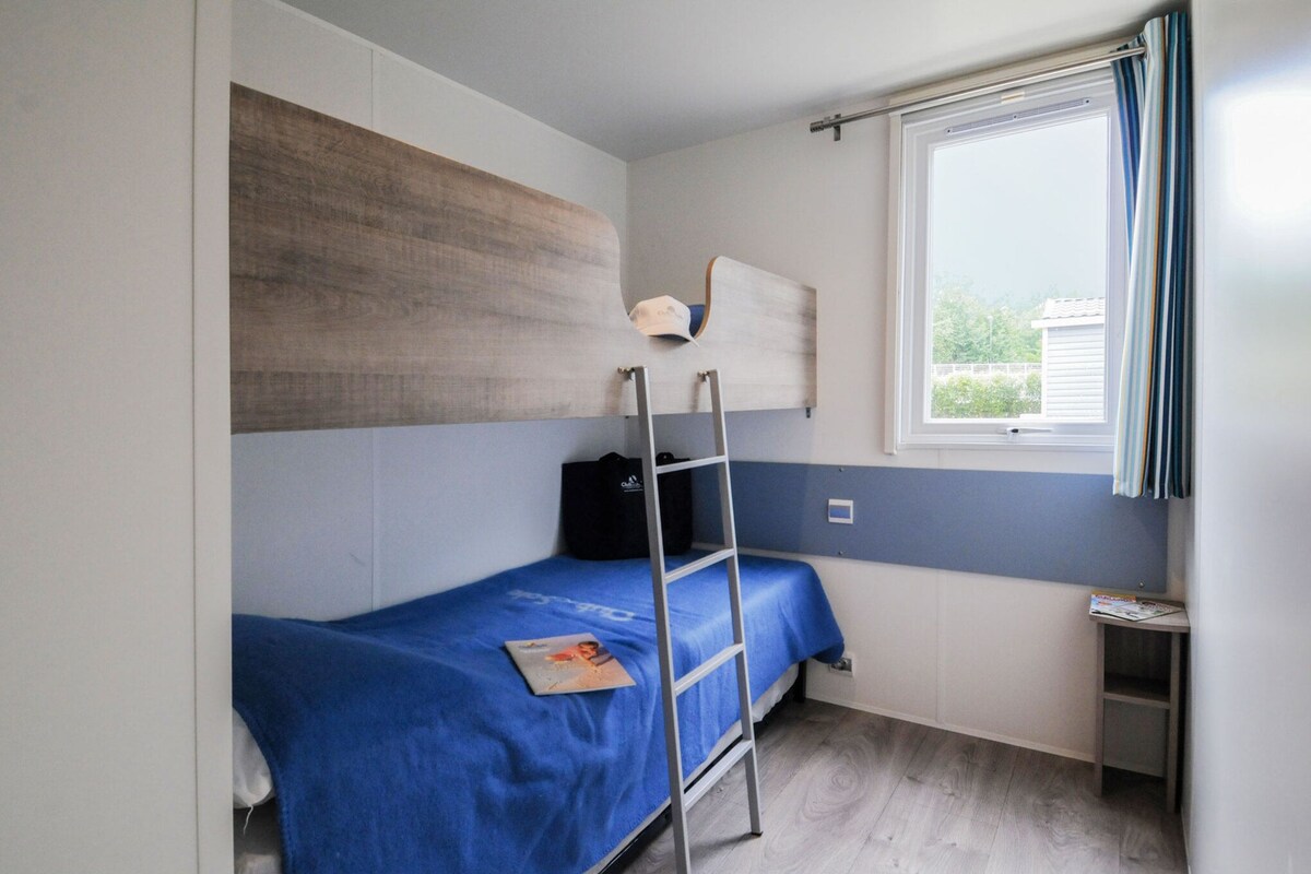 A small, compact room with a bunk bed, a ladder, a window with curtains, and a small table with a magazine.