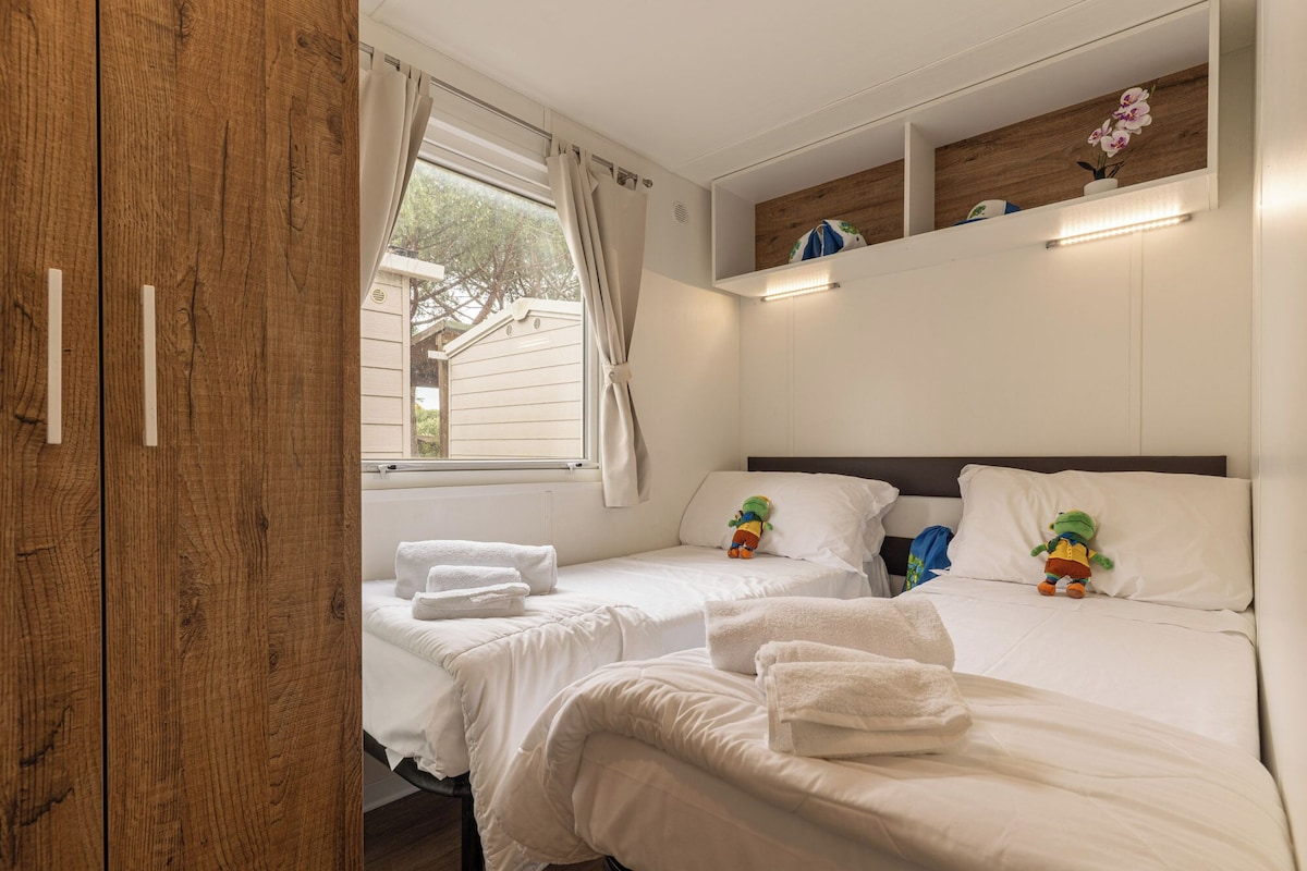 A bedroom with two beds, white bedding, a wooden wardrobe, a window with curtains, and a shelf with decorative items.