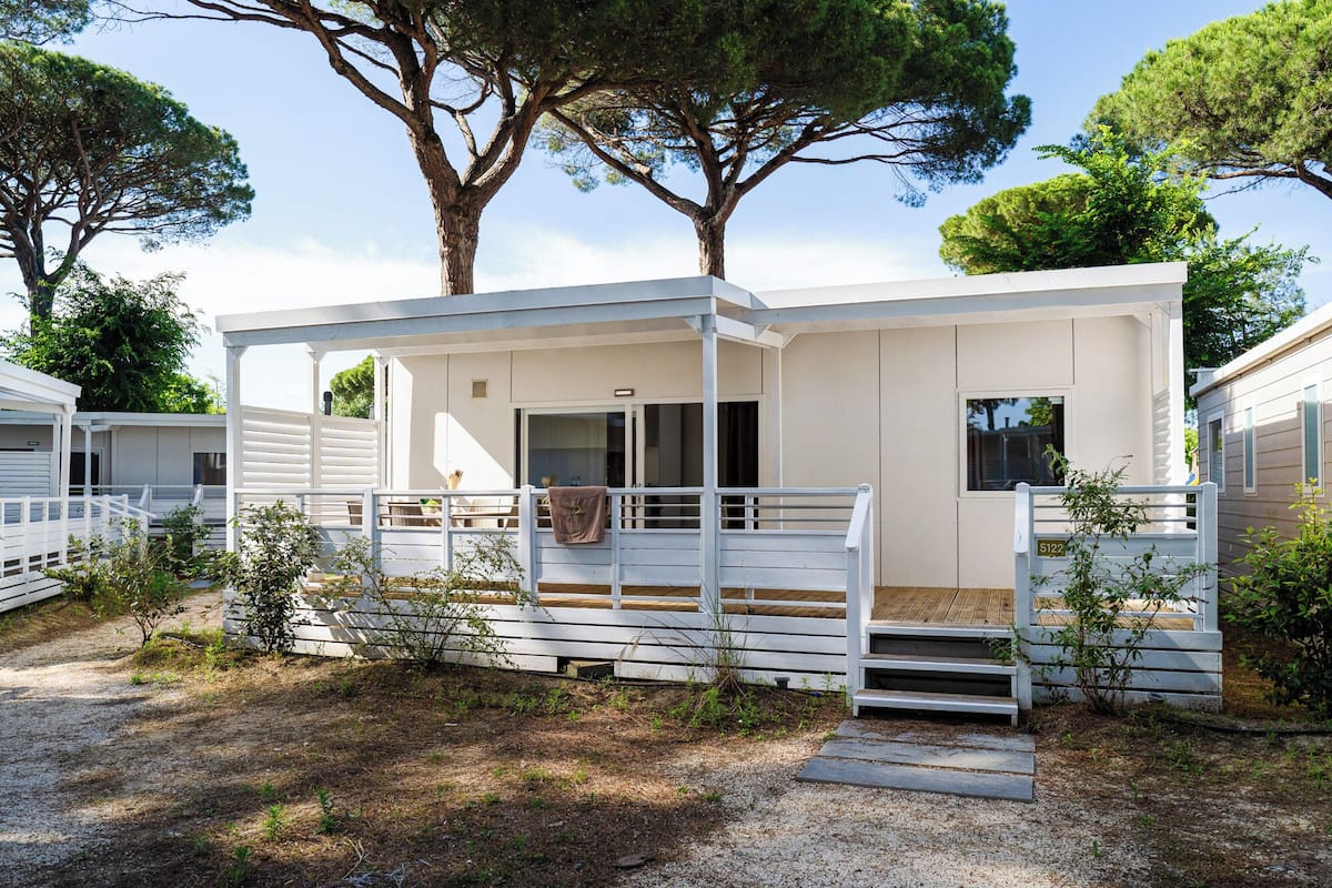 A modern, single-story mobile home with a covered porch, a small outdoor seating area, and a gravel driveway.