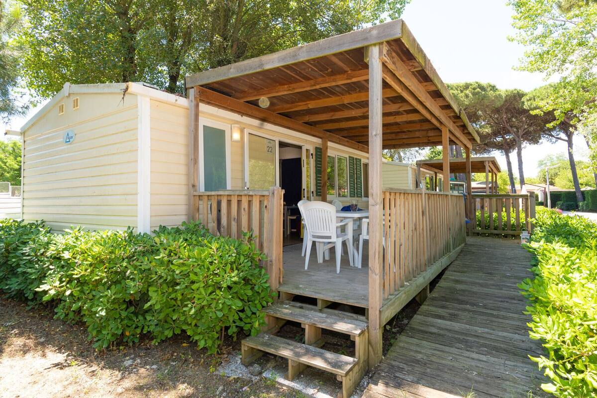 A wooden deck leading to a covered porch with a table and chairs, surrounded by greenery.