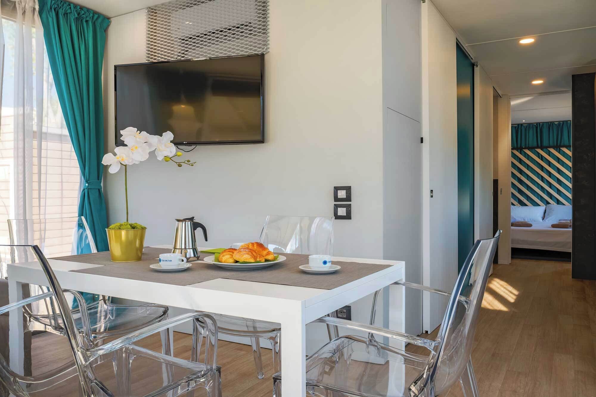 Zona pranzo moderna con tavolo bianco, sedie trasparenti, TV a parete e vista su una camera da letto con testiera a righe.