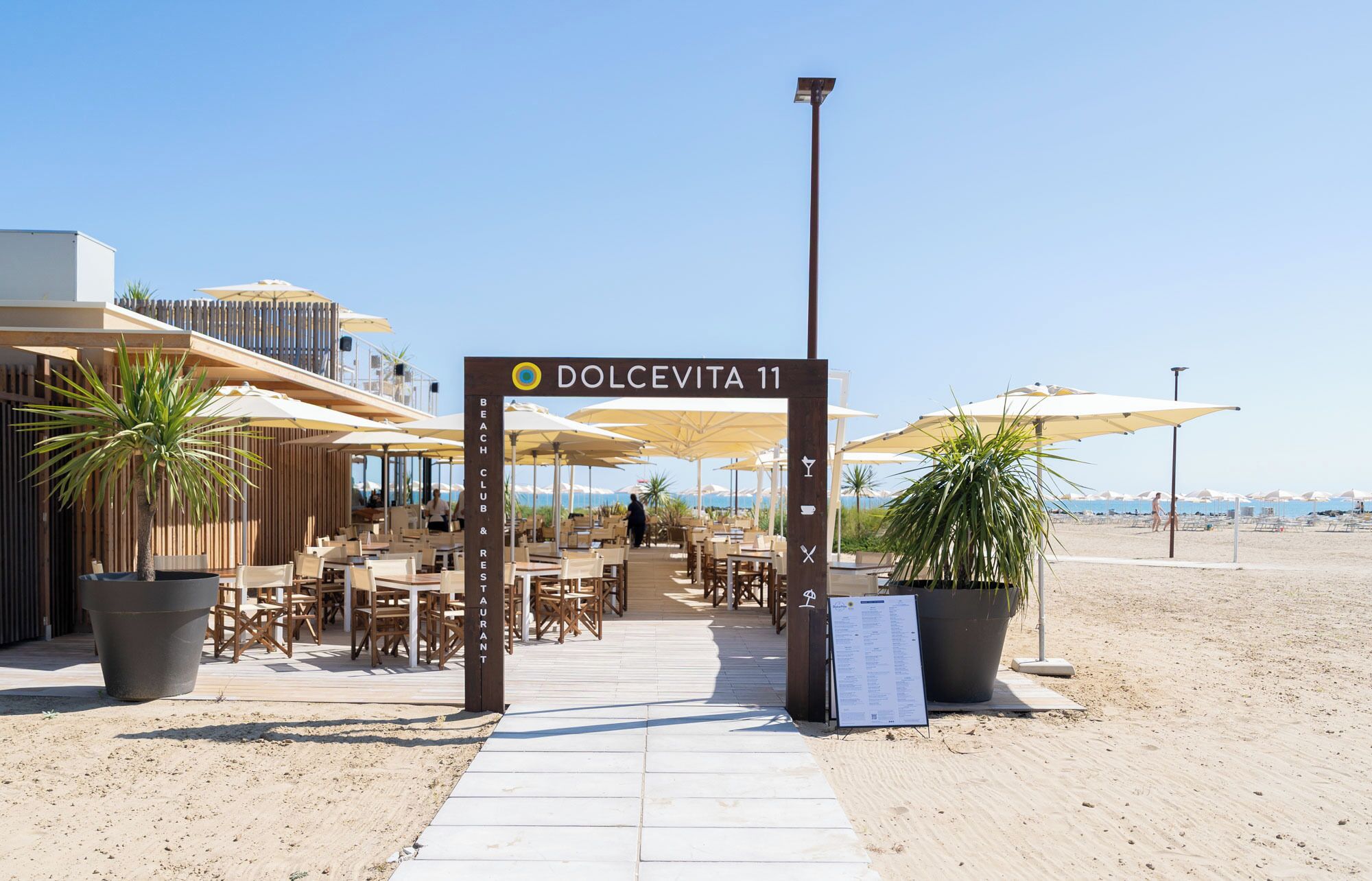 Entree van een strandclub met terras, een bord met 'Dolcevita 11' en een menukaart.