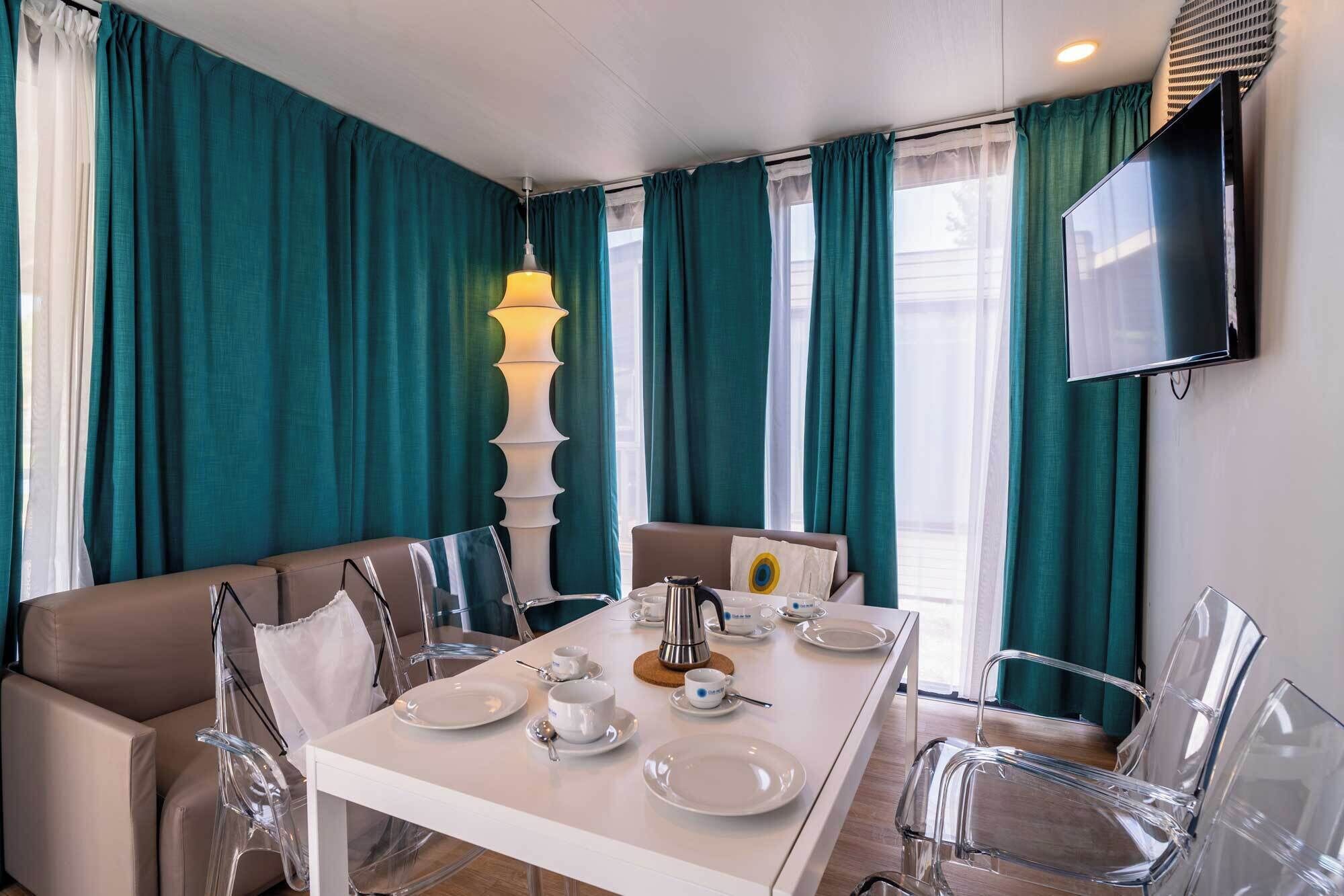 Un coin repas avec une table dressée pour quatre personnes, des chaises transparentes, une cafetière et un téléviseur fixé au mur.