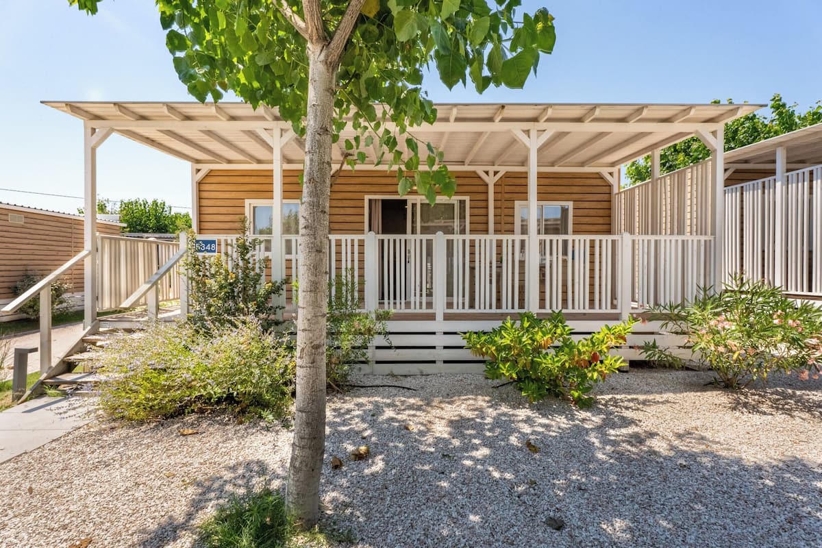 A wooden cabin with a covered porch, gravel ground, and a small tree in the foreground.