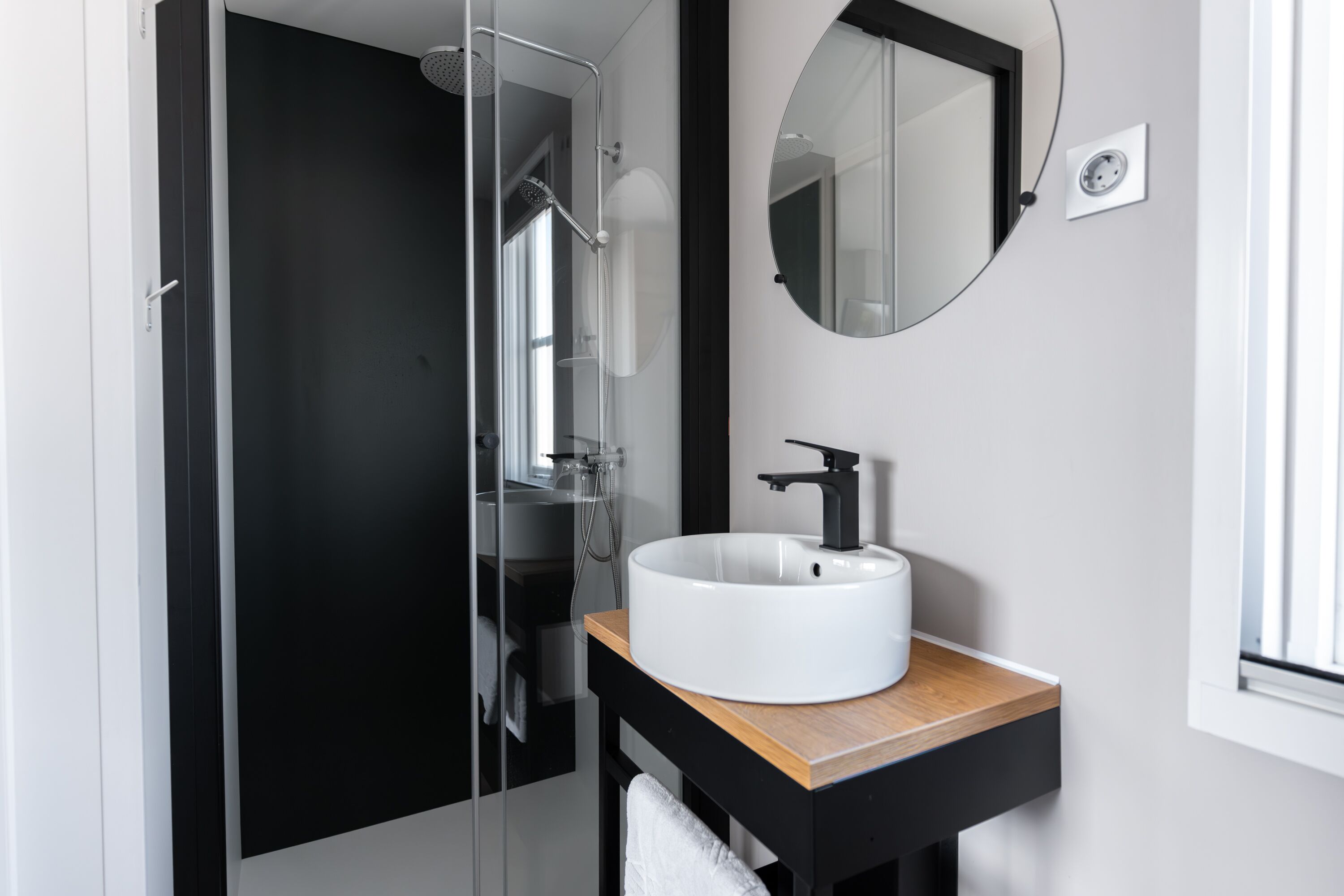Une salle de bain moderne avec une douche à parois de verre, un lavabo blanc doté d’un robinet noir et un plan de travail en bois.