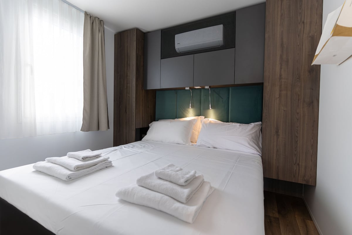 A neatly made bed with folded white towels, a headboard with built-in lighting, and a window with sheer curtains.