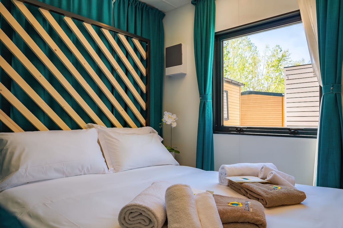 A neatly made bed with white linens and rolled towels, a striped headboard, and a window with curtains.
