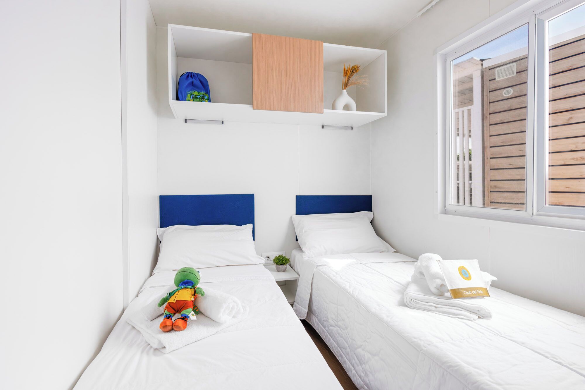 Une chambre d’hôtel avec deux lits, une petite étagère sur laquelle se trouvent un sac et un vase, une fenêtre avec des stores et une serviette munie d’une étiquette.
