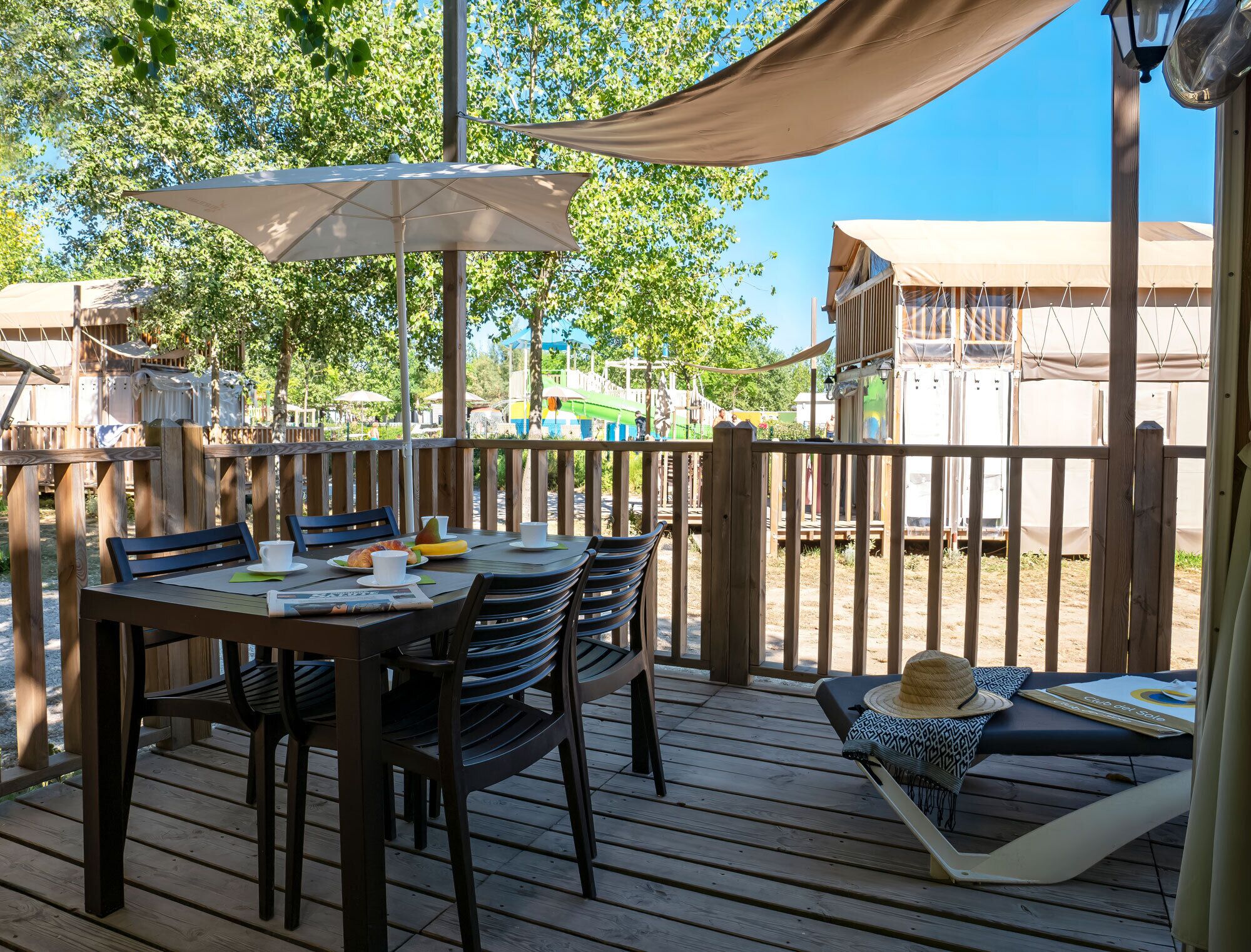 A wooden deck with a table and chairs, a covered area with an umbrella, and a view of a building with a white facade.