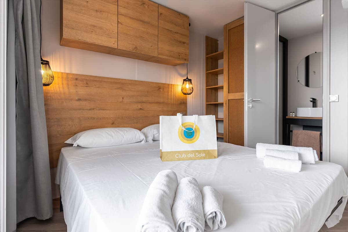 A hotel room with a wooden headboard, a bed with white linens, a towel rack, a wooden wardrobe, and a bathroom with a mirror and sink.