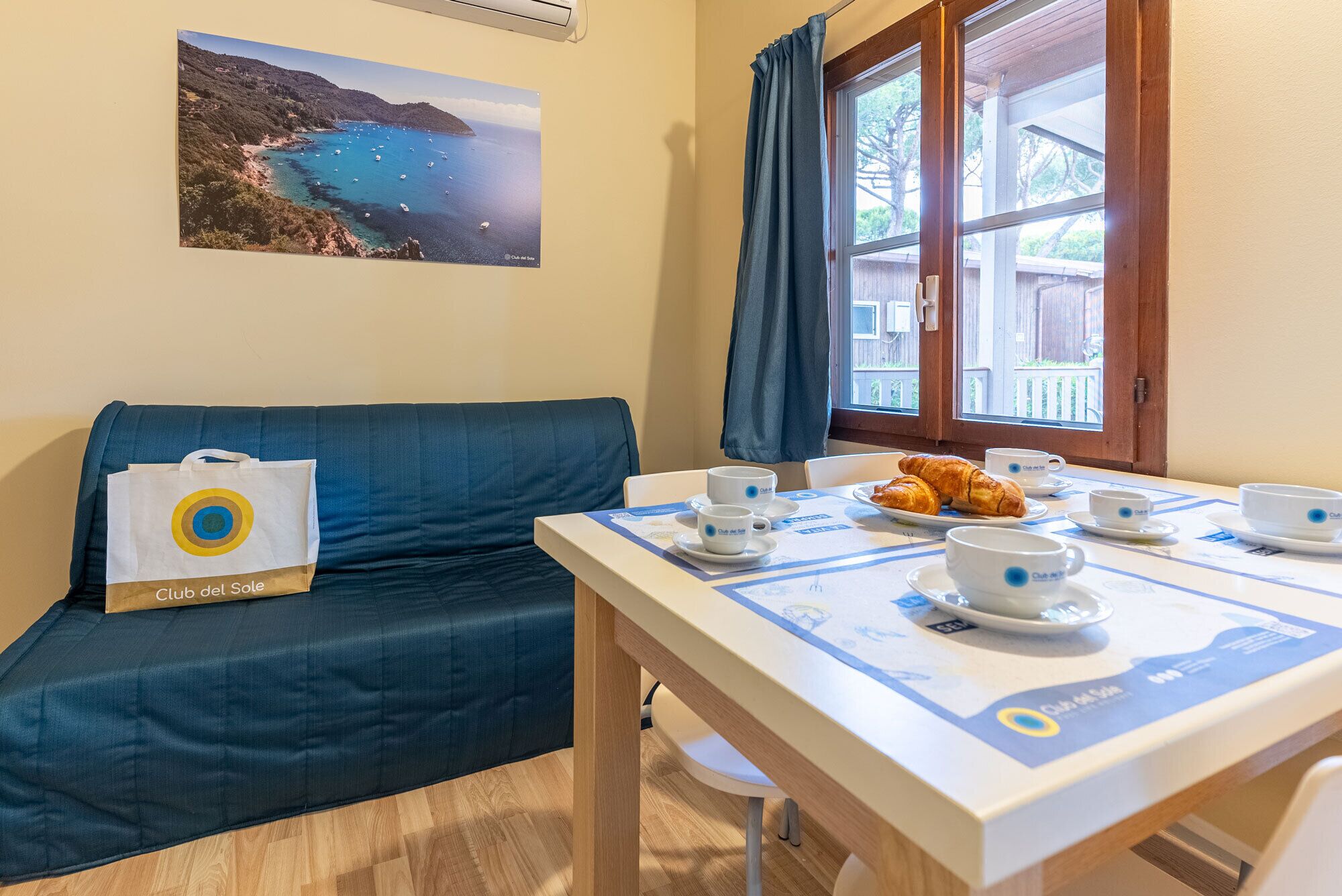 A room with a sofa, a table set for breakfast, a window with curtains, and a picture on the wall.