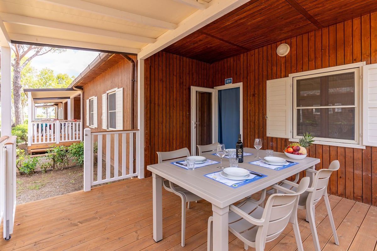 Un espace extérieur abrité, aménagé pour les repas, avec une table pour deux personnes, des chaises blanches et une terrasse en bois.